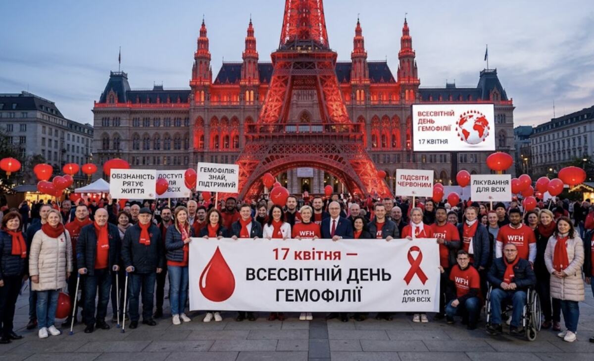 Учасники акції тримають великий банер "17 квітня — Всесвітній день гемофілії" на тлі підсвіченої червоним будівлі