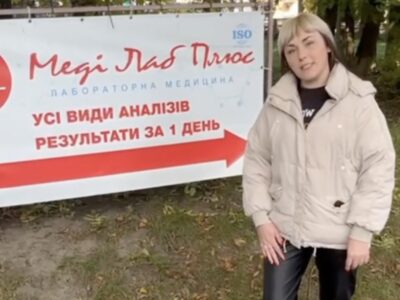Жінка вказує на рекламний банер медичної лабораторії Меді Лаб Плюс біля Хмельницької районної лікарні на Львівському шосе