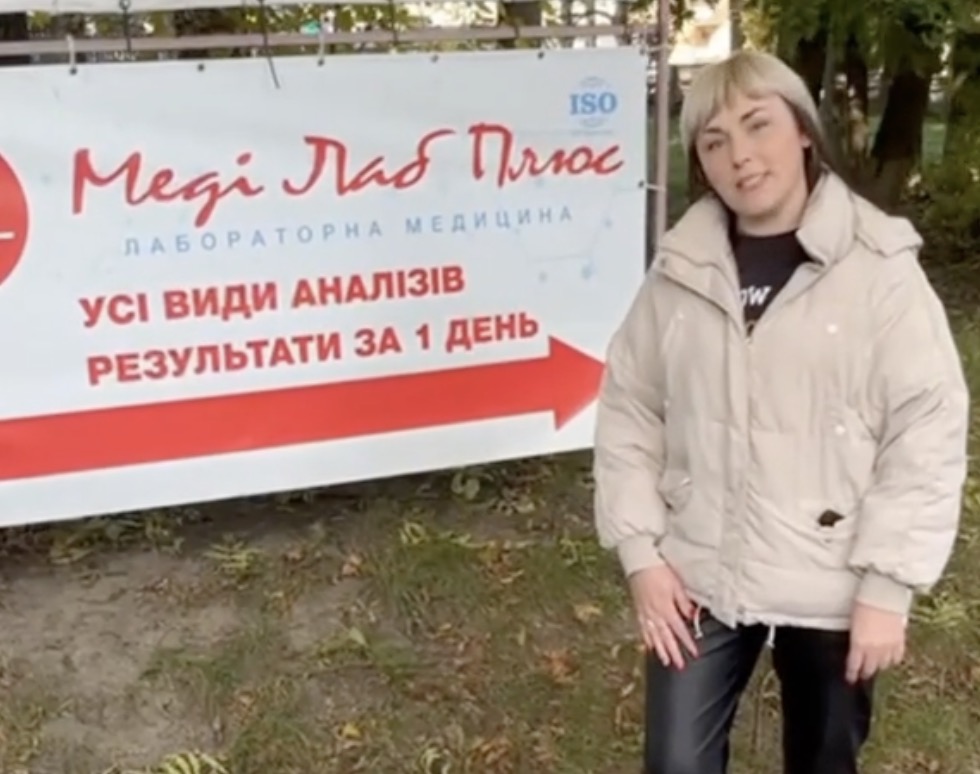 Жінка вказує на рекламний банер медичної лабораторії Меді Лаб Плюс біля Хмельницької районної лікарні на Львівському шосе